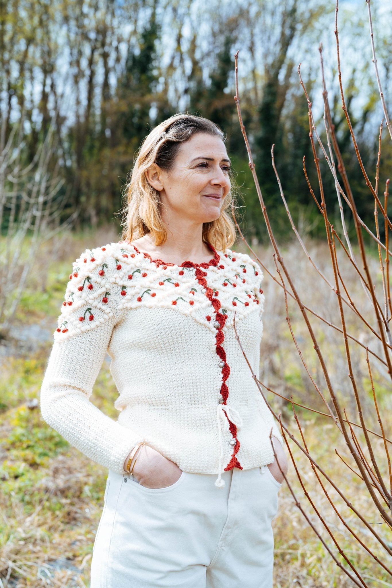 Woman wearing Ernestine cream handknitted cardigan with cherry embroidery in natural outdoor setting.